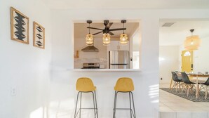 A modern kitchen with a built-in oven, a stainless steel refrigerator, and pendant lights. There are two yellow bar stools in front of the kitchen. - ✧VIEWS✧ 6min to Joshua Tree walk to bars + firepit  (Twentynine Palms)
