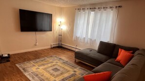 En stue med et vægmonteret fladskærms-tv, en gulvlampe, en sofa med orange puder og et mønstret tæppe.