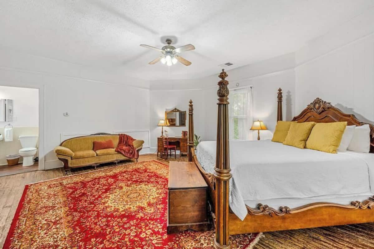 A bedroom with a four-poster bed, a red patterned rug, a wooden chest of drawers, a sofa, and a ceiling fan.