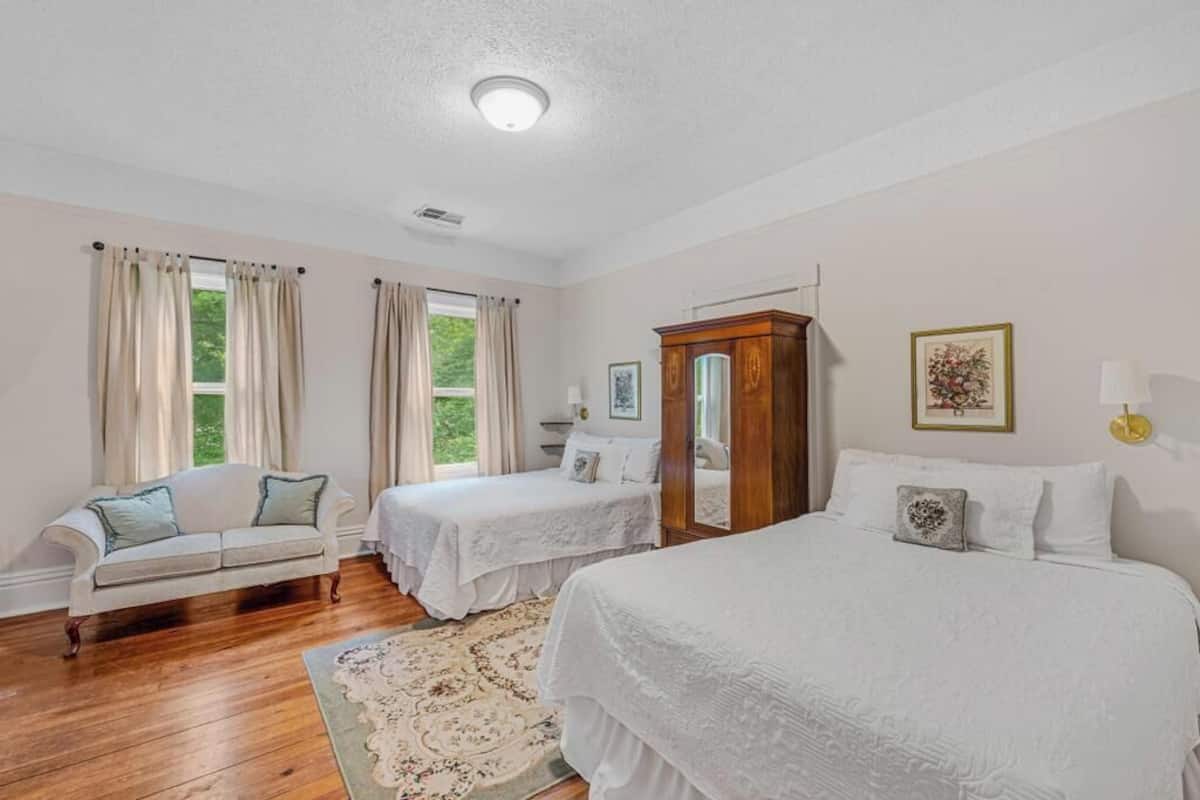 A bedroom with two beds, a wooden wardrobe, a white sofa, and a large window with curtains.