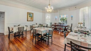 A dining area with wooden flooring, white walls, and a chandelier. Tables are set for dining with chairs.