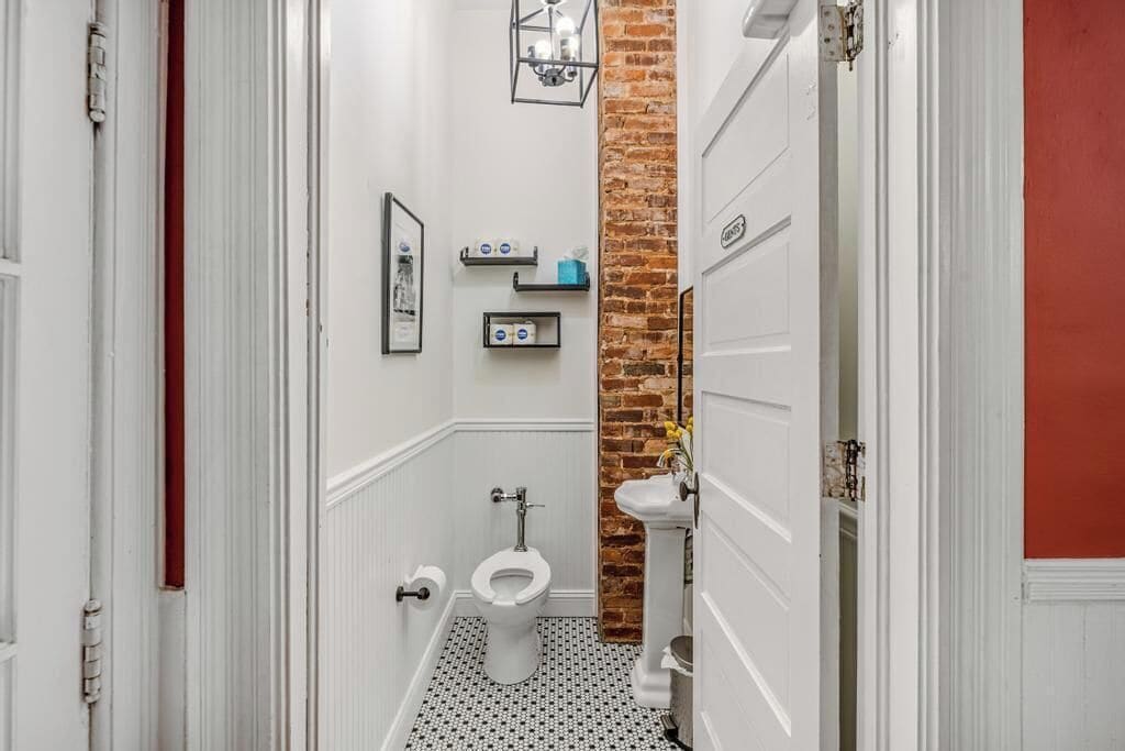 A narrow bathroom with a white toilet, a small sink, and a brick accent wall.