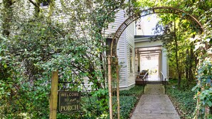 A wooden sign welcoming visitors to a porch, surrounded by lush greenery and a pathway leading to a house with a covered porch. - The Veranda Historic Inn (Senoia)