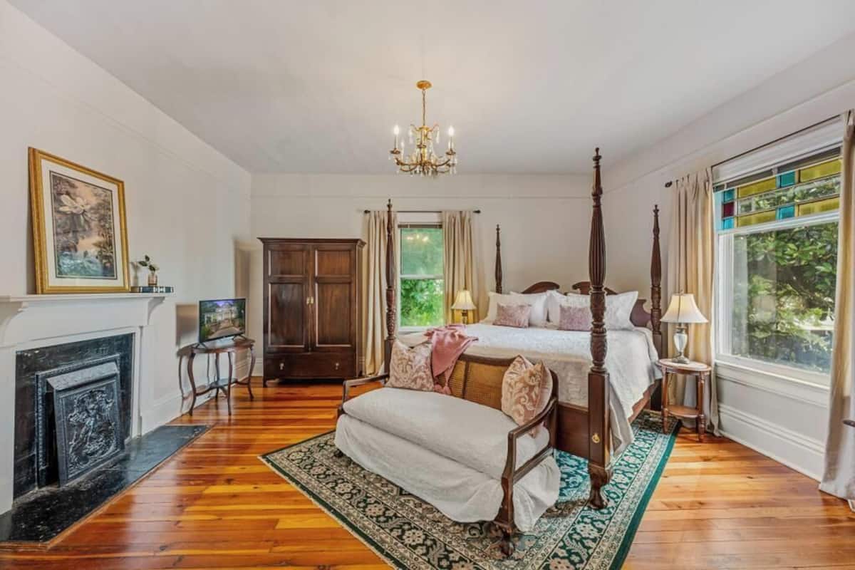 A spacious bedroom with a four-poster bed, a fireplace, and a large window with curtains.