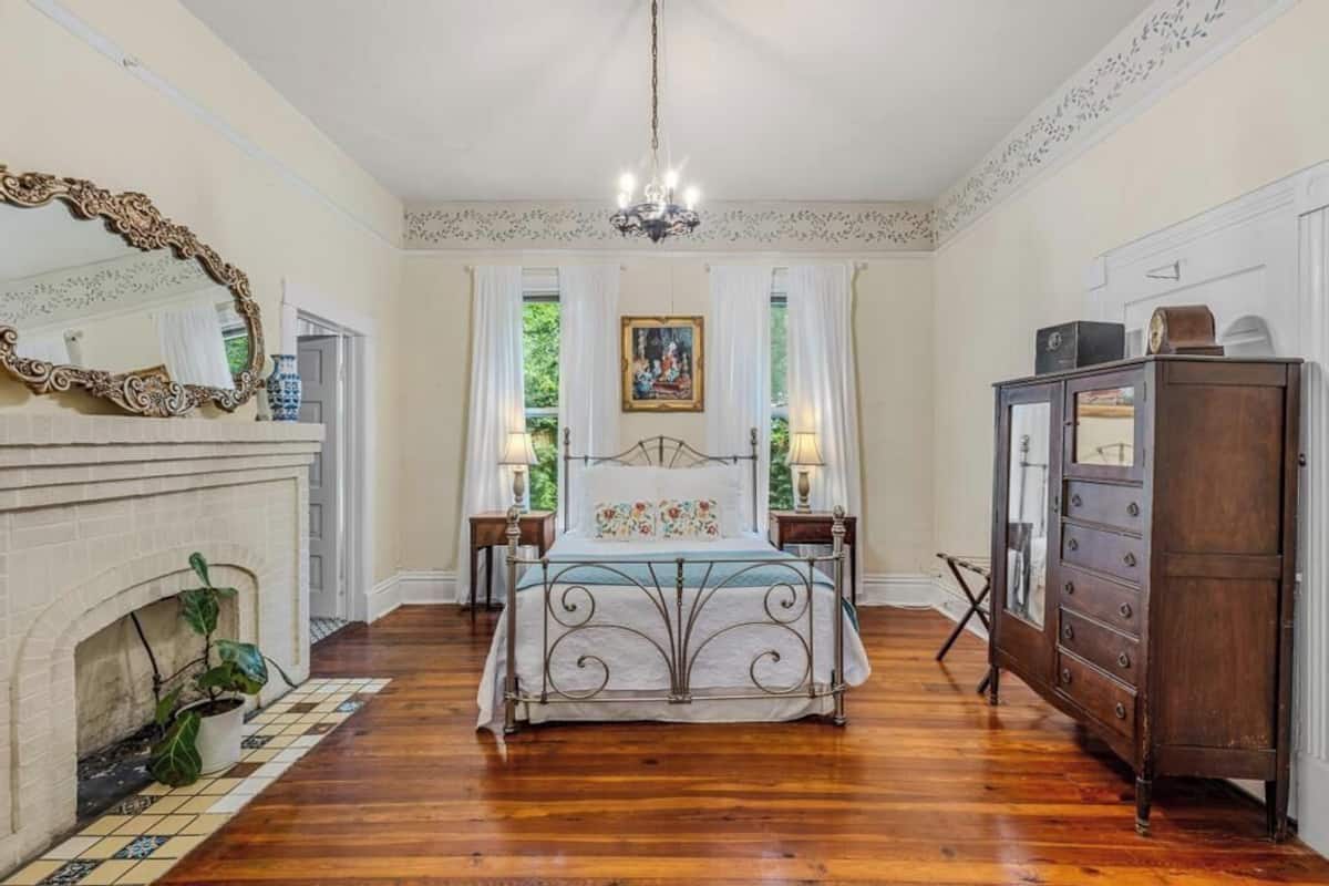 A bedroom with a bed, a dresser, a mirror, a fireplace, and a chandelier.