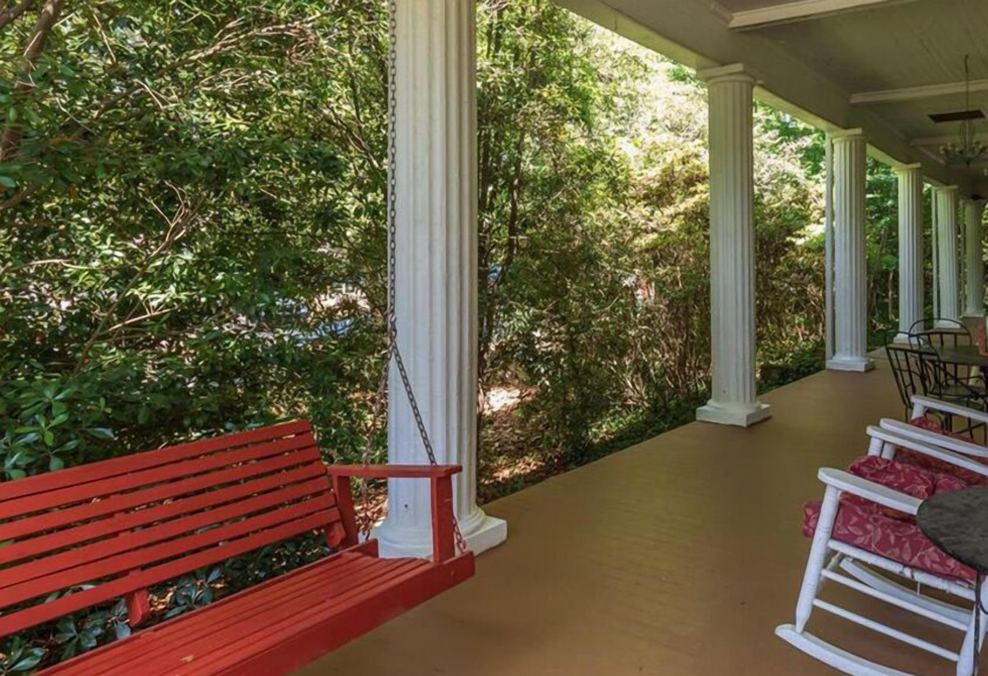 A covered porch with a red bench and white rocking chairs, surrounded by greenery.