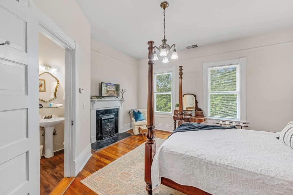 A bedroom with a four-poster bed, a fireplace, a large window, and a door leading to a bathroom.
