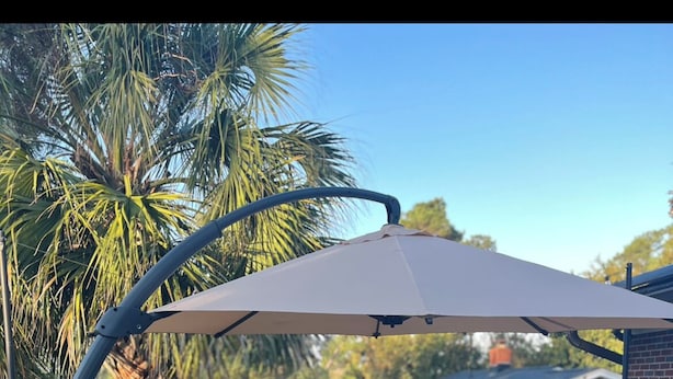 Eine Holzterrasse mit gepolsterten Sitzgelegenheiten im Freien, eine Pergola mit Dach und im Hintergrund Palmen und ein Haus.