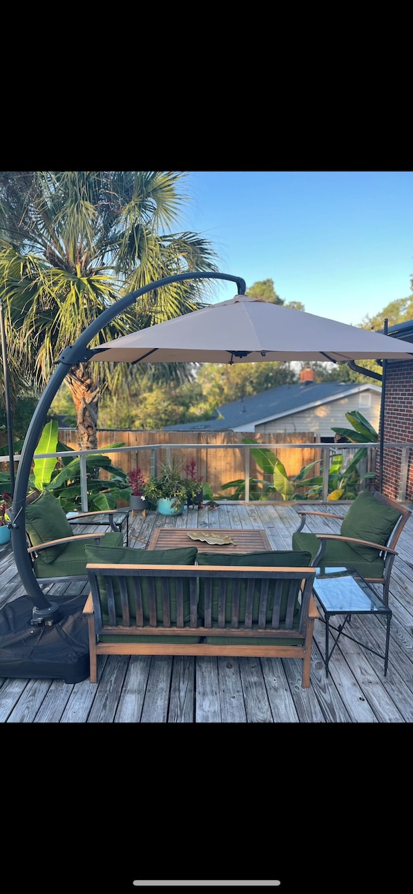 A wooden deck with a cushioned outdoor seating area, a pergola with a canopy, and a backdrop of palm trees and a house.