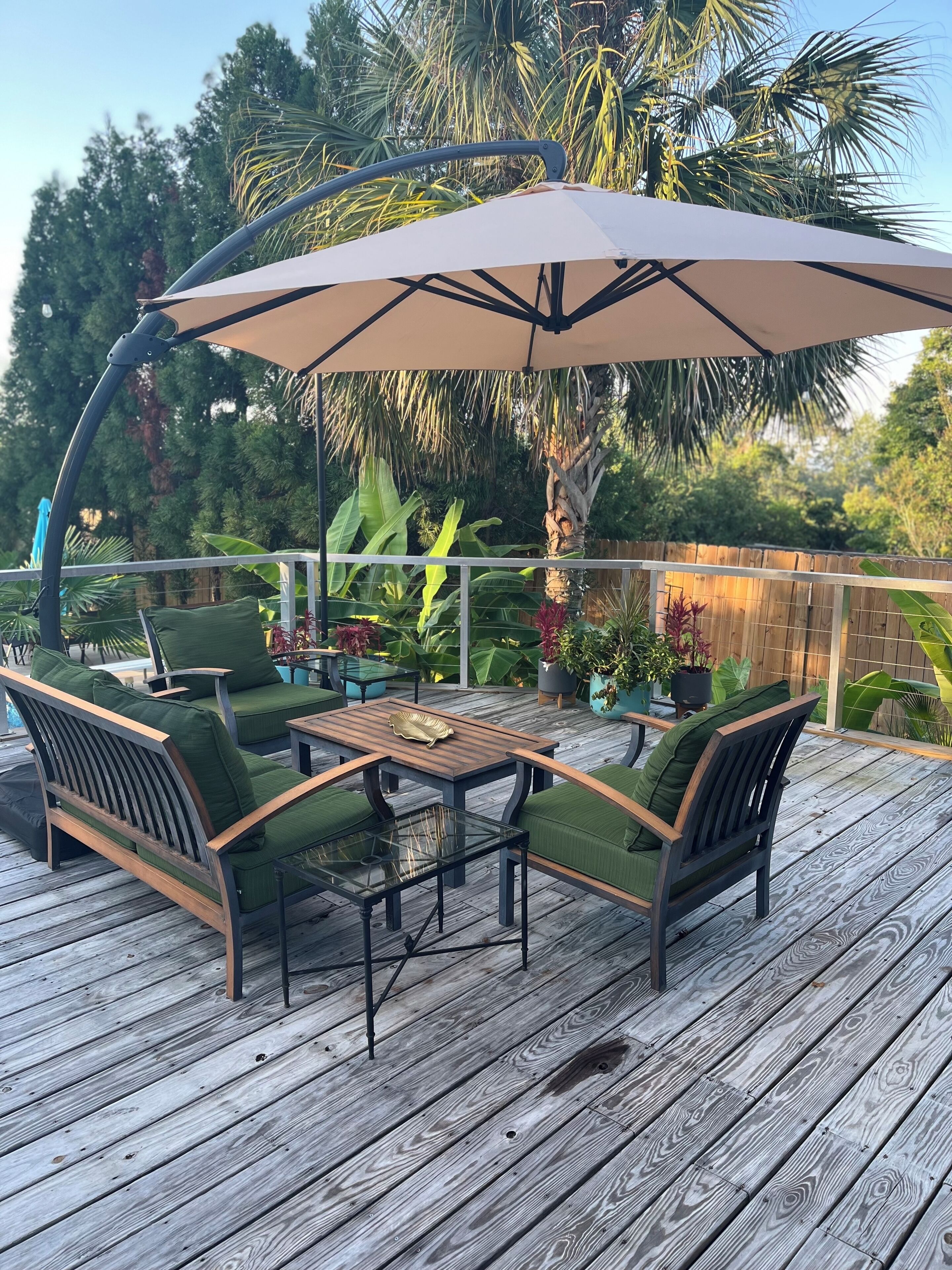 An outdoor patio with a wooden deck, green cushioned chairs, a glass-top table, and a large umbrella.