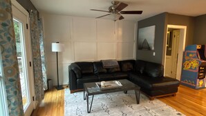 Ein Wohnzimmer mit einem schwarzen Ledersofa, einem Couchtisch, einem gemusterten Teppich, einem Deckenventilator und einem großen Fenster mit Vorhängen.