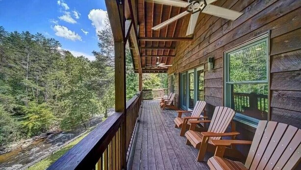 A wooden deck with Adirondack chairs, overlooking a forested area and a river.