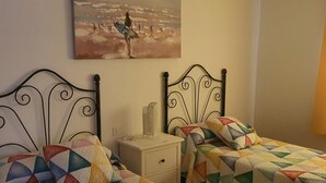 A bedroom with two beds, each with a colorful geometric-patterned bedspread. There is a nightstand with a lamp and a small decorative item between the beds. A painting hangs on the wall above the beds. - APARTAMENTO FAMILIAR (Caleta de Famara)