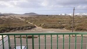 A view from a balcony with a green railing, overlooking a desert landscape with sparse vegetation and distant buildings. - APARTAMENTO FAMILIAR (Caleta de Famara)