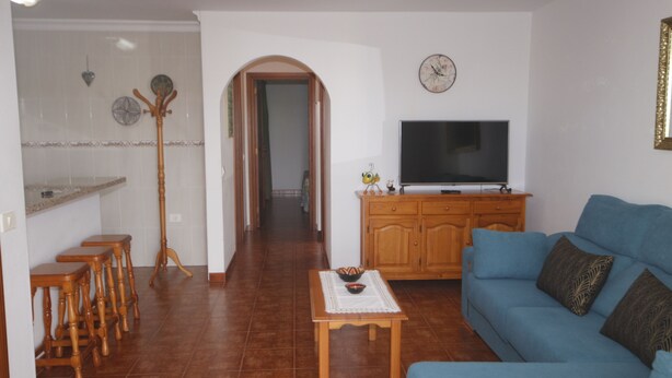 A living room with a blue sofa, wooden coffee table, and a television on a wooden cabinet. There are two wooden stools and a coat rack with hats. - APARTAMENTO FAMILIAR (Caleta de Famara)