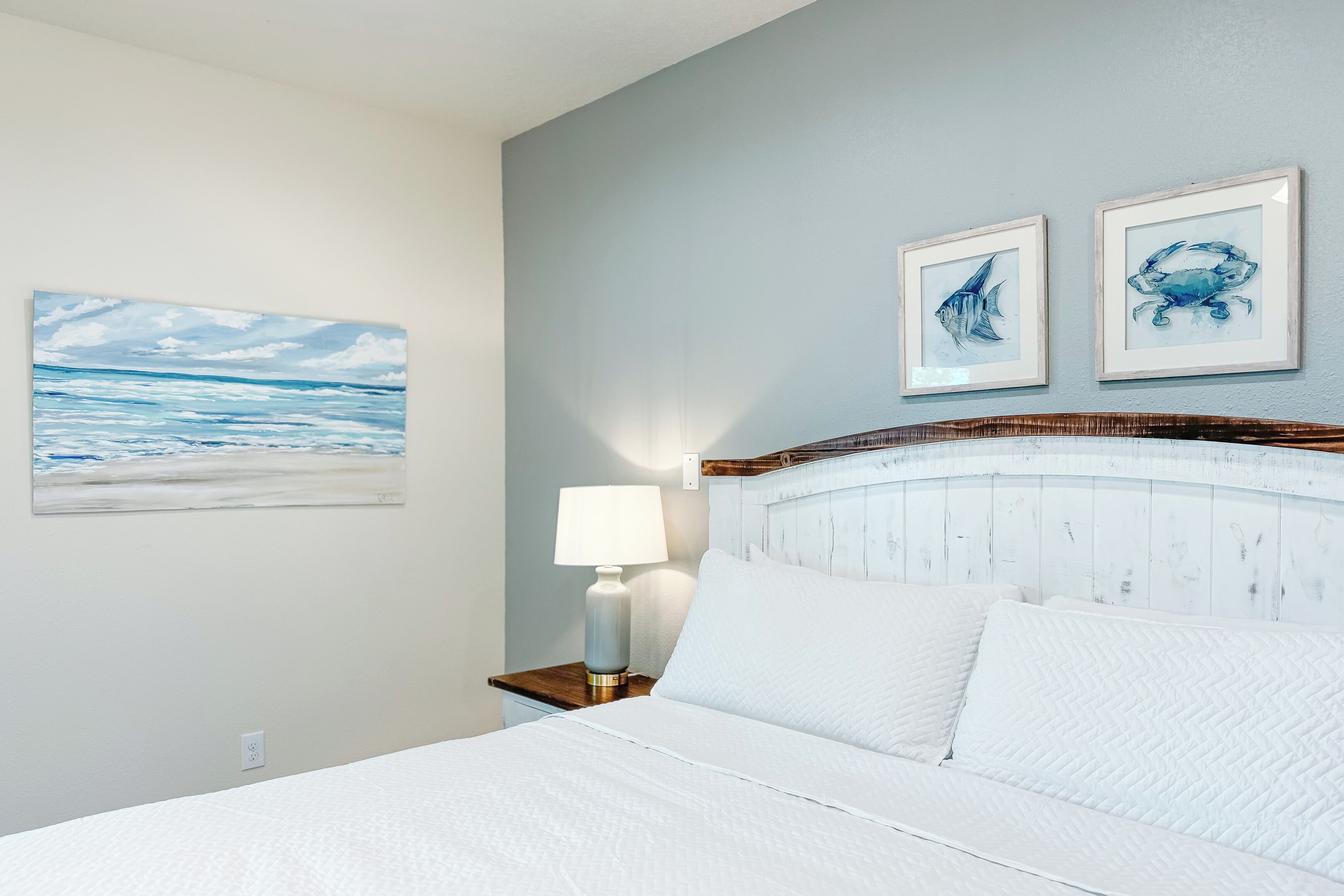 Una cama bien hecha con ropa de cama blanca, un cabecero de madera y una mesita de noche con una lámpara. Hay dos cuadros enmarcados en la pared sobre la cama.