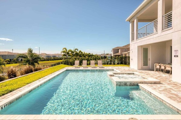 A swimming pool with a clear blue water feature, surrounded by a landscaped garden and a building with outdoor seating. - Colorful Getaway with Home Theater at Encore (Kissimmee)