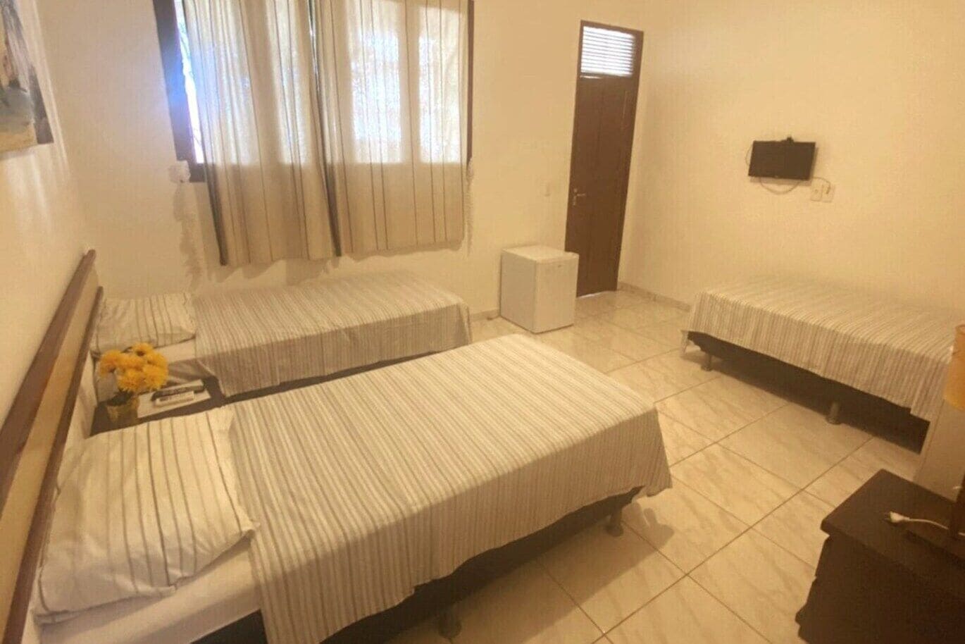 A hotel room with three single beds, a small bedside table with a lamp, a window with curtains, a door, and a wall-mounted TV.