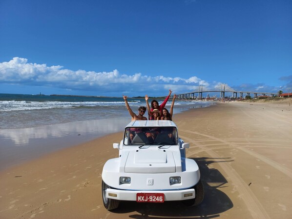 A white off-road vehicle on a sandy beach with a bridge and cityscape in the background.