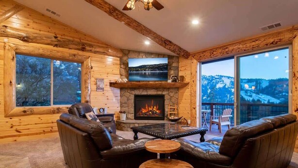 A log cabin interior with a fireplace, leather sofas, a stone fireplace surround, and a large window overlooking a snowy landscape. - Sky Ridge. Movie Room. Games. Views. Spa. BBQ. (Big Bear Lake)