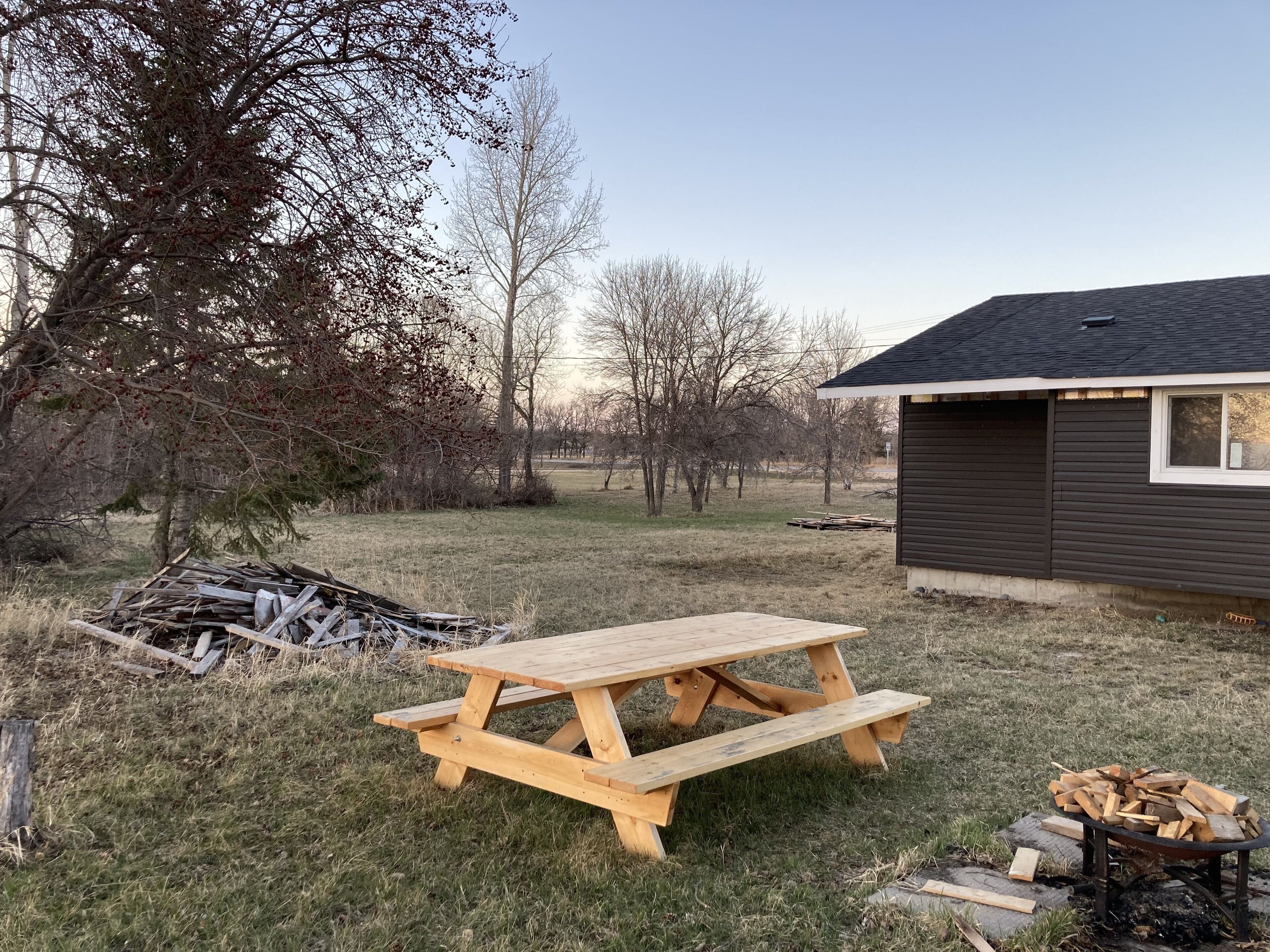 Une table de pique-nique en bois se trouve dans une cour verdoyante, à proximité d’une pile de bois de chauffage.
