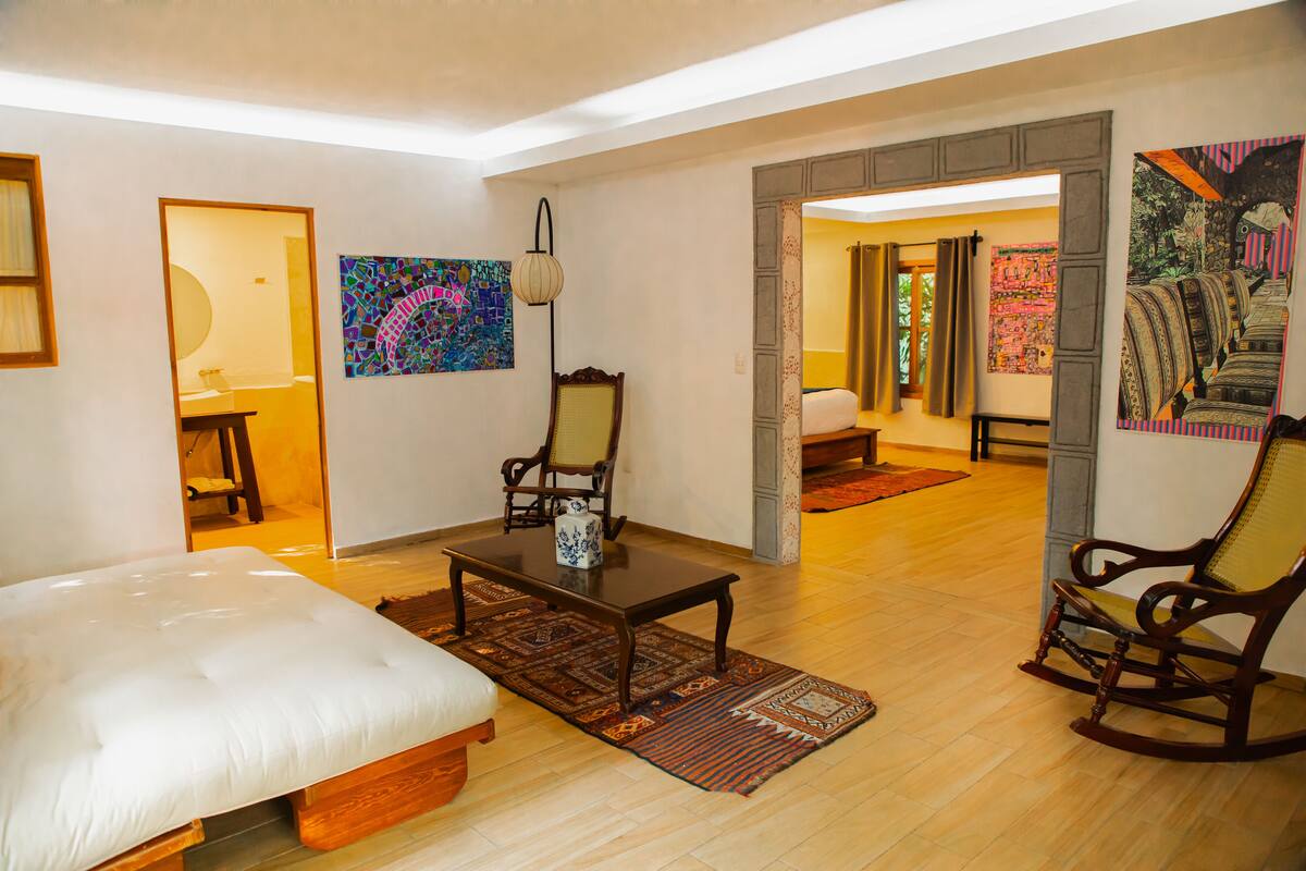 Amplia sala de estar con una cama grande, una mesa de centro de madera y dos mecedoras. Se observan obras de arte coloridas en las paredes y una puerta que da a otra habitación.