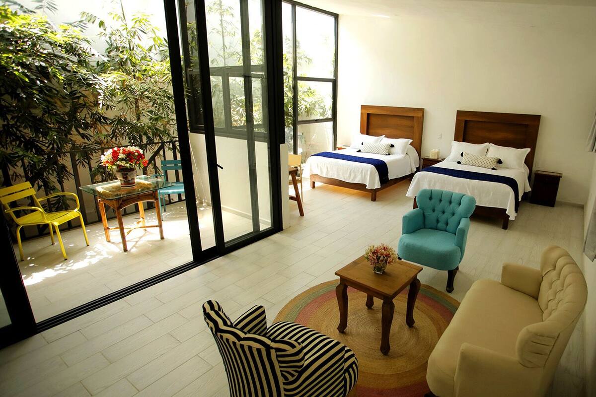 Habitación de hotel con un amplio ventanal corredero que da a un balcón, una cama con cabecera de madera, un sillón azul, un sofá color beige y una mesita con un florero.