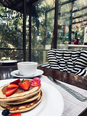 Desayuno con tortitas, fresas y arándanos en un plato, una taza de café y una mesa con vistas a los árboles a través de amplias ventanas.