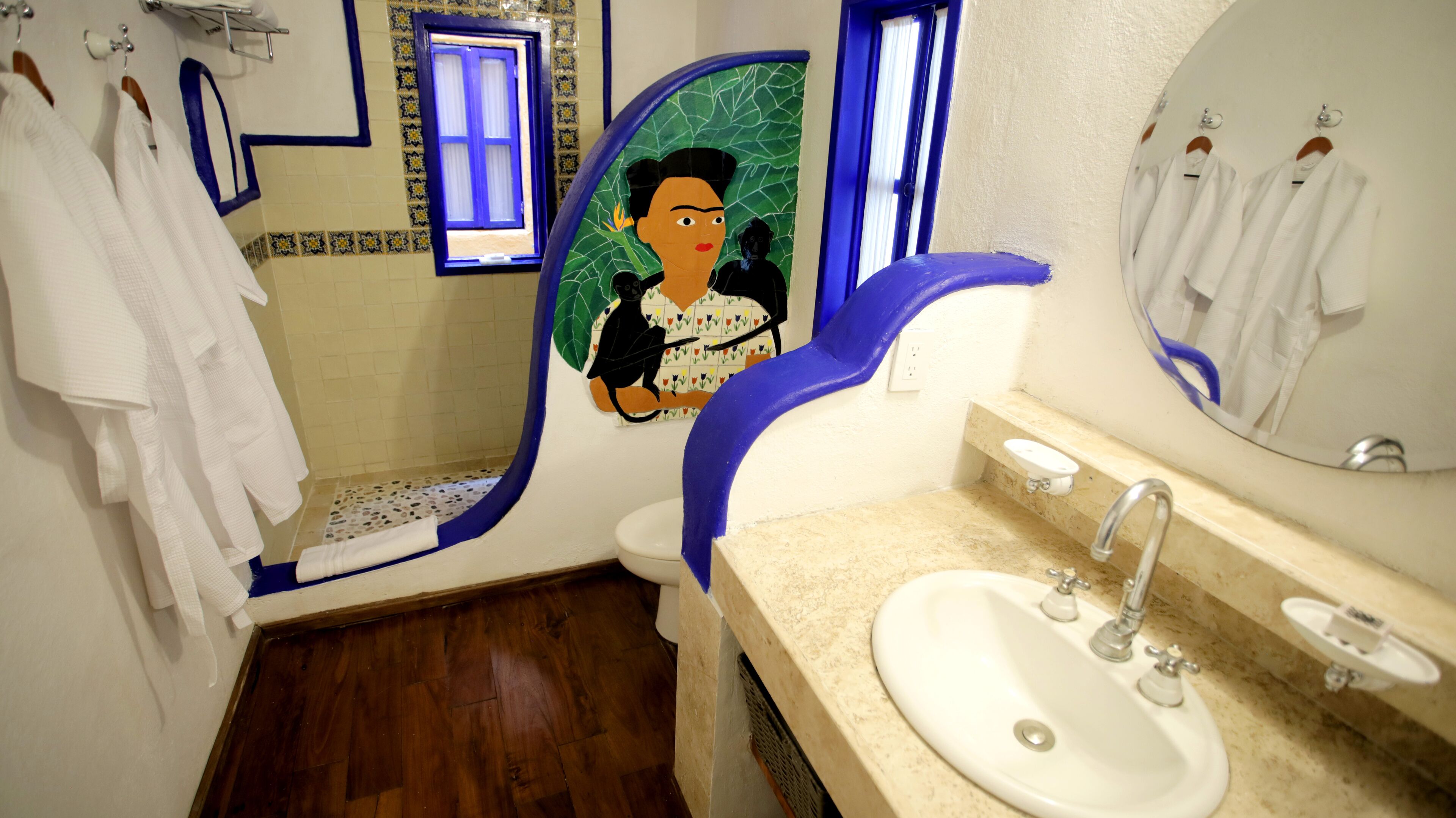 Un baño con lavamanos, espejo y retrete. Hay toallas colgadas en la pared y una ventana con detalles azules.