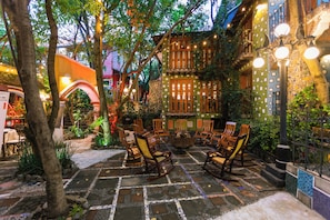 Une terrasse extérieure avec des chaises en bois et un foyer, entourée de verdure et dotée d’un sol en pierre.