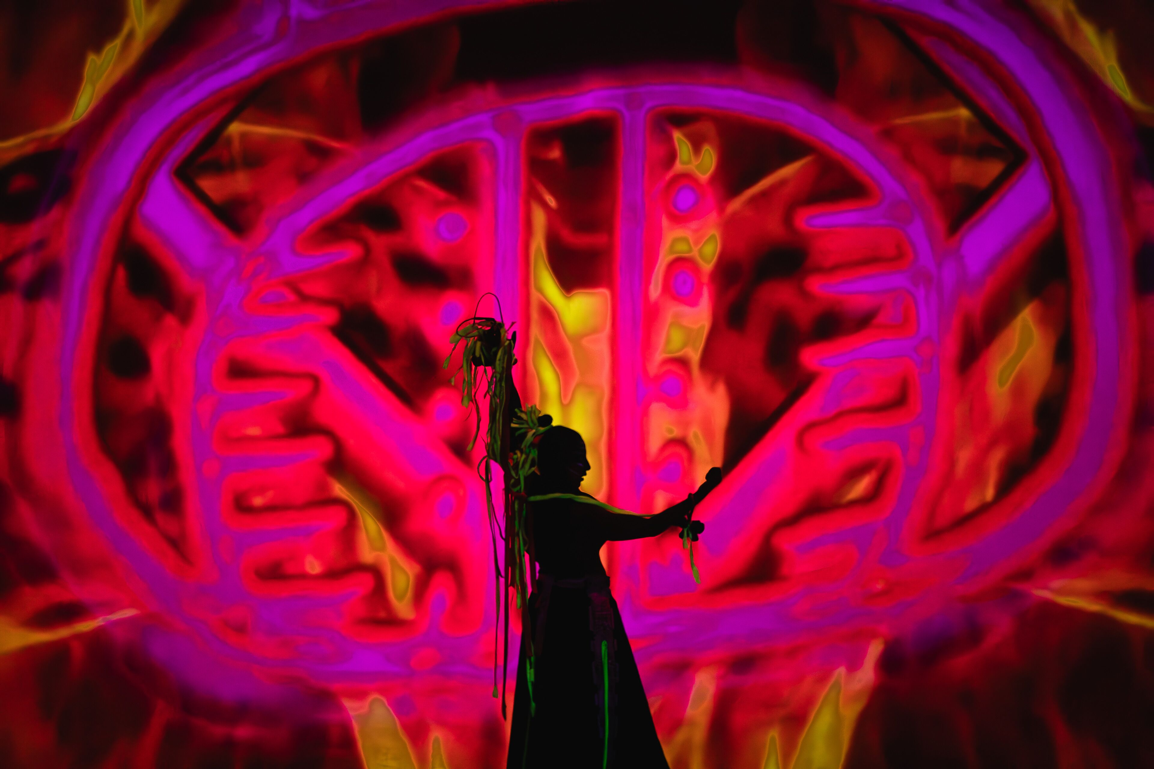 A person in a long dress holding a staff, standing in front of a large, glowing, circular structure.