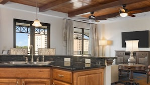 A hotel room with a kitchen area, wooden cabinets, a dining table, a chair, a ceiling fan, and a large window with a view of buildings.