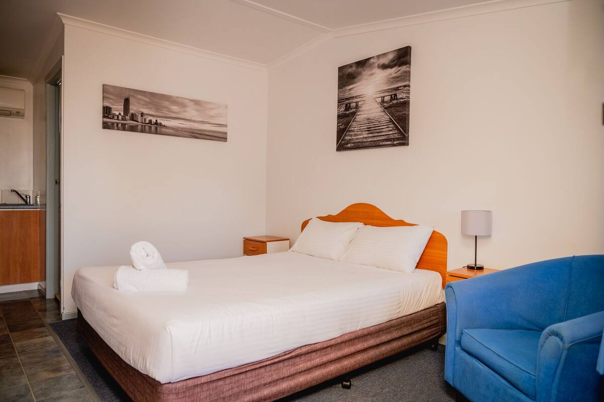 Een hotelkamer met een bed, een blauwe fauteuil, een nachtkastje en twee kunstwerken aan de muur.