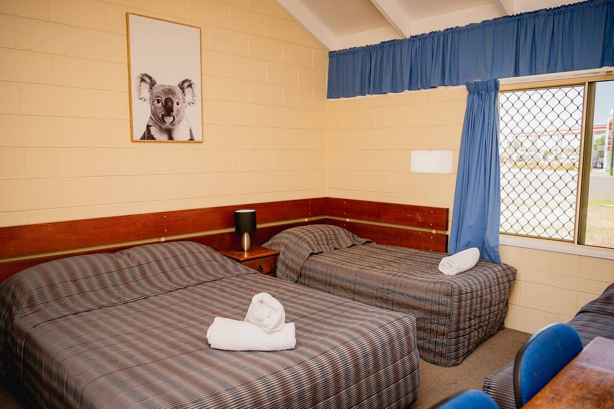 Hotelkamer met twee bedden, een houten hoofdbord, een lijst met afbeelding van een koala, een raam met blauwe gordijnen en een tafel met een stoel.