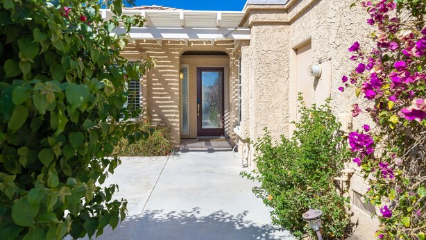 A building with a concrete pathway leading to a door, surrounded by greenery and flowering plants. - Stylish 3 bedroom condo w/ office & 3 pools (Palm Desert)