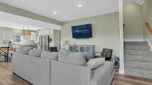 Ein modernes Wohnzimmer mit einem grauen Ecksofa, einem Essbereich mit Holztisch und einer Küche mit weißen Schränken und einem eingebauten Backofen.