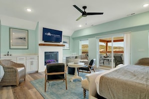 A hotel room with a bed, a dining area with chairs, a fireplace, and a balcony with outdoor furniture.