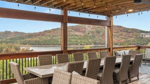 Eine Terrassengruppe im Freien mit Korbmöbeln, einem Holztisch und Blick auf einen See sowie bewaldete Hügel.
