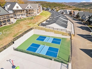 A tennis court with a surrounding fence, adjacent to a row of houses with sloped roofs. - Enjoy a Private Pool with Panoramic Views, Elevator Access, Private Golf Cart & Free Tickets! (Hollister)