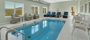 An indoor swimming pool with lounge chairs and tables, large windows, and a view of the outdoors.