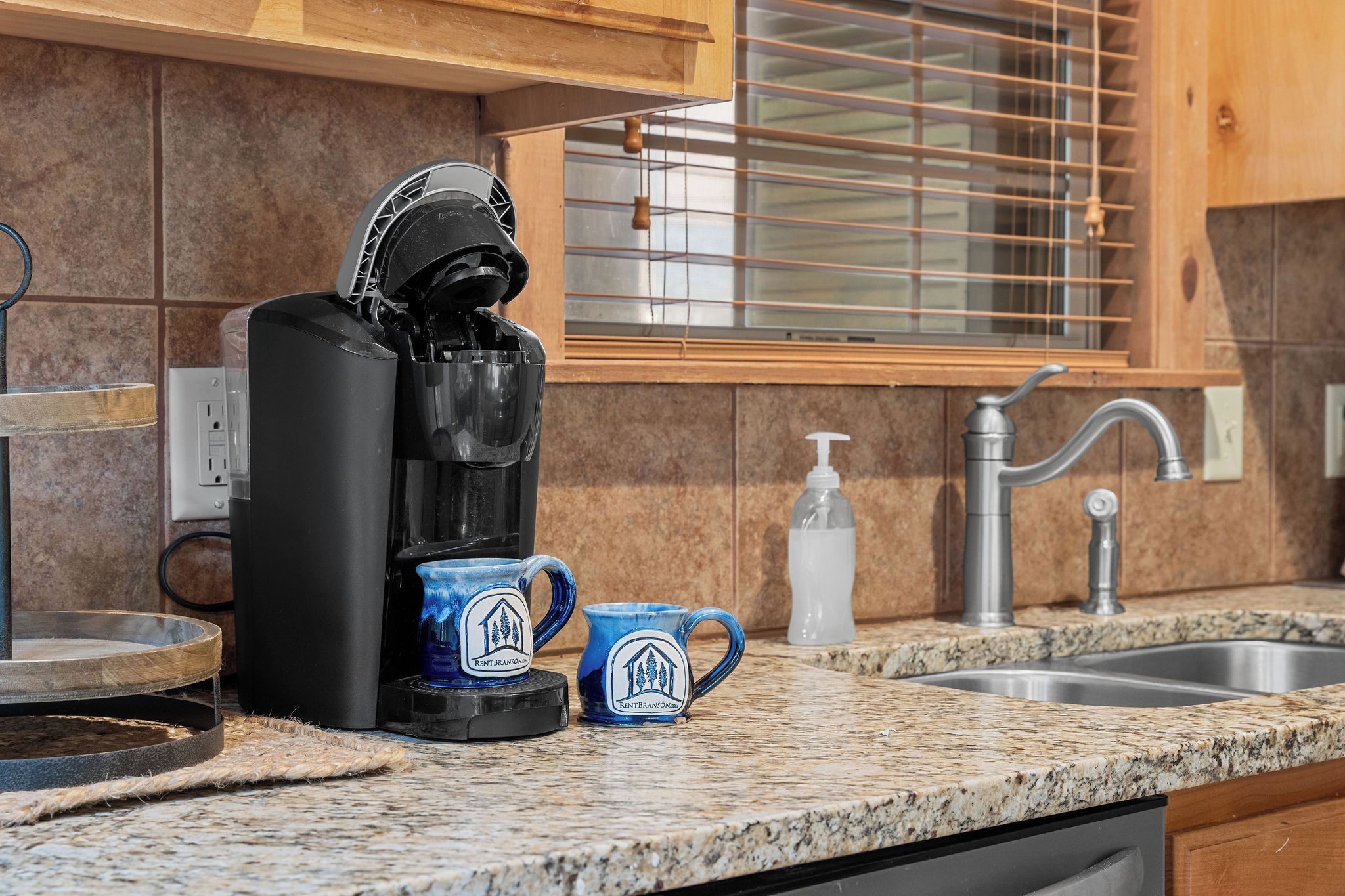 A kitchen with a coffee maker, two mugs, a soap dispenser, and a stainless steel sink.