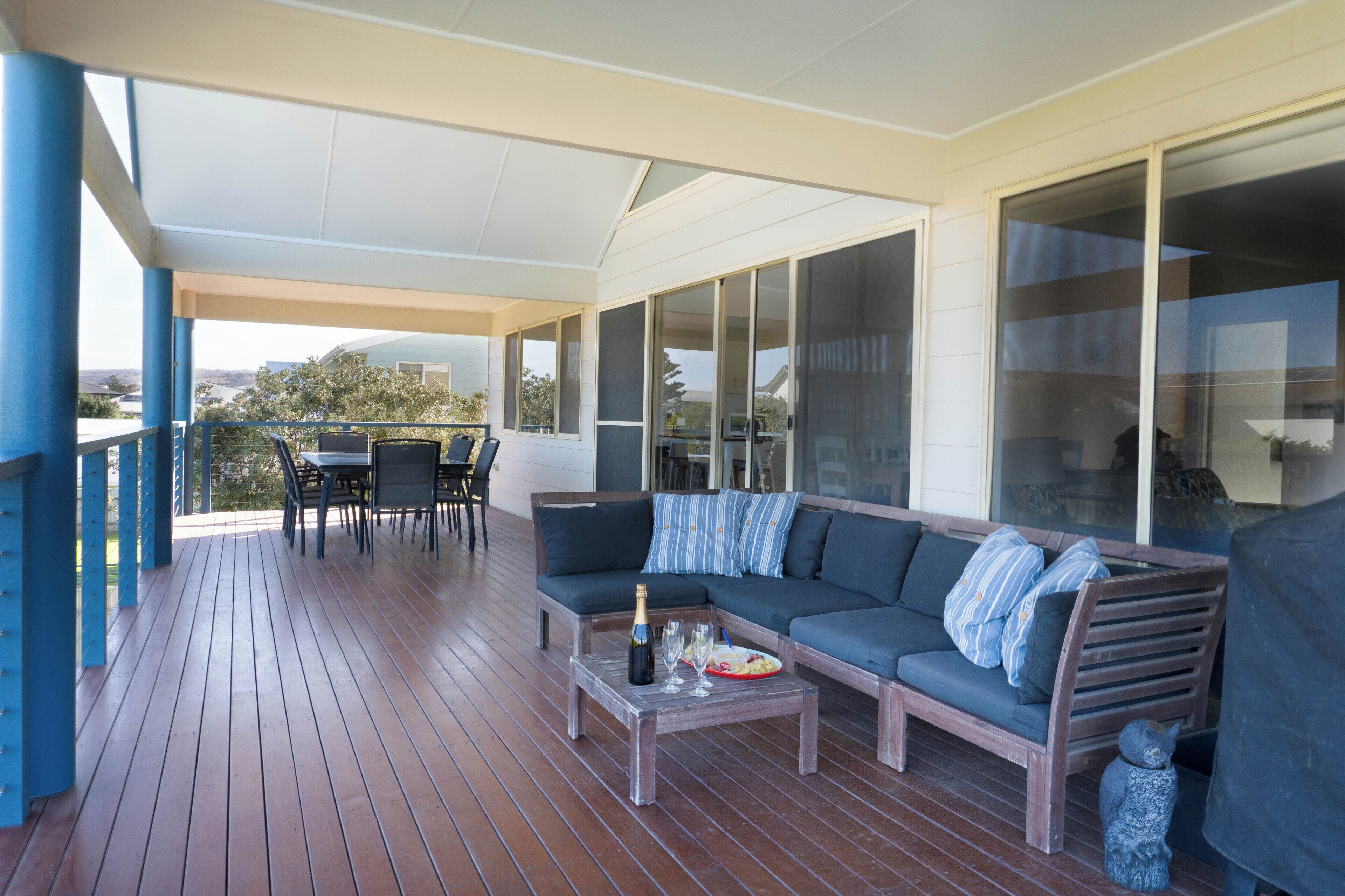 A covered outdoor seating area with a wooden deck, a sofa, a table with a bottle and glasses, and a chair.