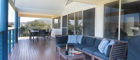 A covered outdoor seating area with a wooden deck, a sofa, a table with a bottle and glasses, and a chair.