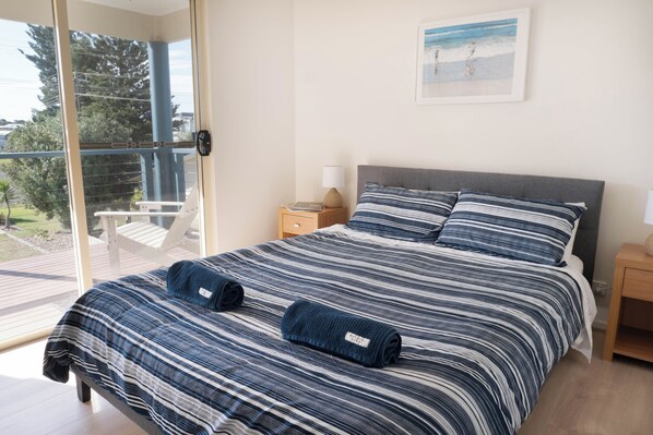 A bedroom with a large bed, two pillows, a nightstand, a lamp, and a window with a view of a balcony and trees. - Two Blue Doors | Coastal Escape, Middleton SA (Middleton)