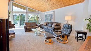 A modern living room with a wooden ceiling, leather sofas, a glass coffee table, and a view through large windows. - Vacation for Families with Stunning View & Pool (Peachland)