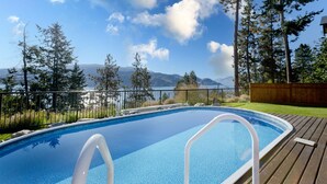 A swimming pool with a wooden deck, surrounded by trees and a view of mountains and a lake. - Vacation for Families with Stunning View & Pool (Peachland)