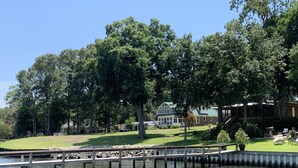 A riverside view with a wooden dock extending into the water, surrounded by lush greenery and a large house with a green roof. - Waterfront King Beds Pet Friendly Deck Home with Dock & Views. (Eutawville)