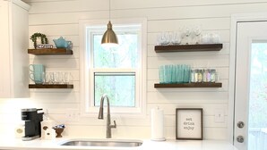 Una cocina con armarios blancos, lavavajillas de acero inoxidable, cafetera y estantes con cristalería y frascos.