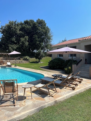 Une piscine entourée de chaises longues et de parasols, elle-même bordée d’une terrasse en pierre et d’une maison dont le toit est recouvert de tuiles.