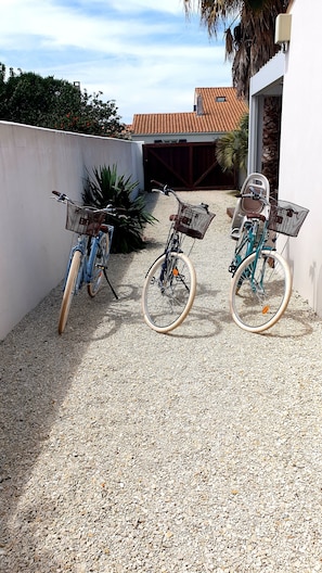 Three bicycles parked on a gravel surface, with a white wall and a building with a tiled roof in the background.